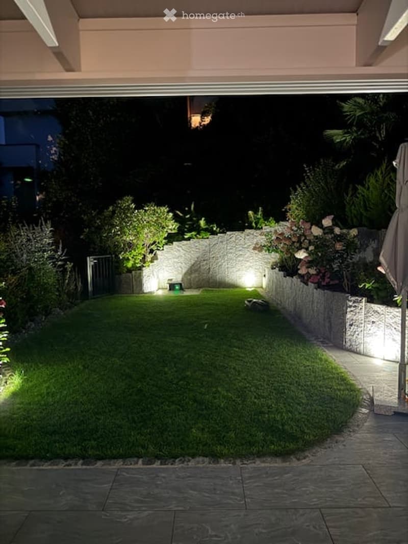 landscaped garden with green lawn, illuminated by spotlights, surrounded by low stone walls and diverse plants