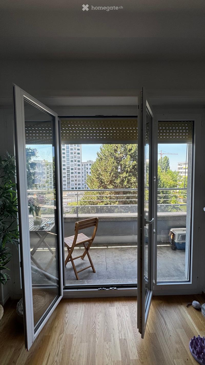Balcony with a wooden chair and small table, overlooking a cityscape with trees and buildings.