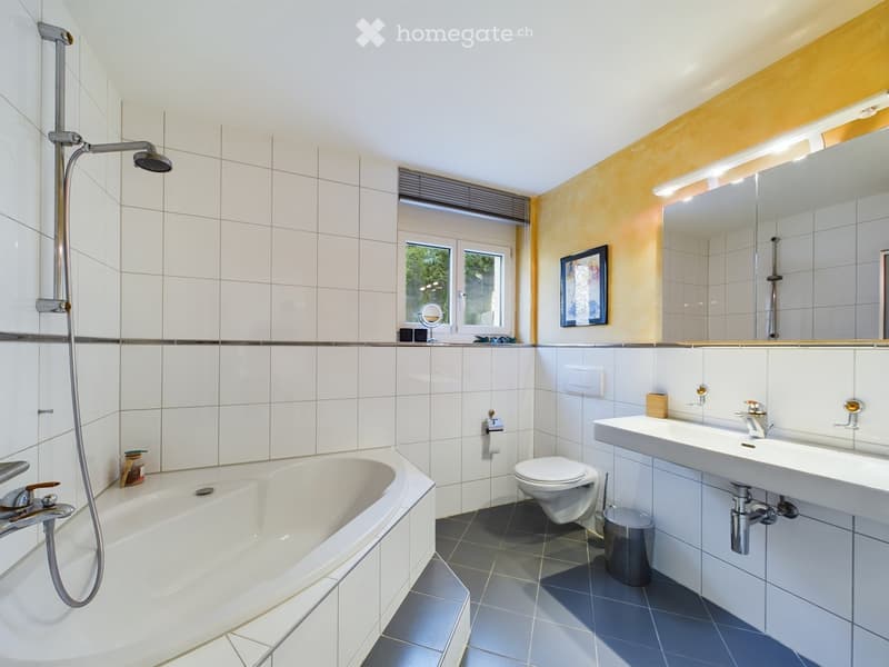 Modern bathroom with white tiled walls, a bathtub, toilet, sink, mirror, and a showerhead. Features a window with blinds and a framed picture on the wall.