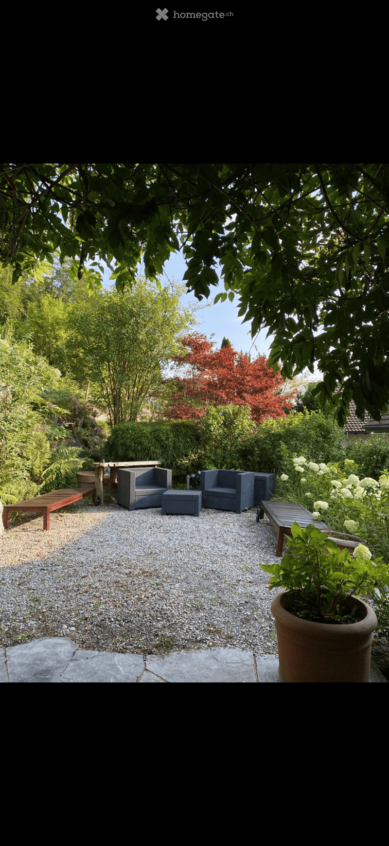 A well-maintained garden with gravel ground, featuring outdoor seating including chairs, a table, a stool, and a bench, surrounded by lush greenery and trees.