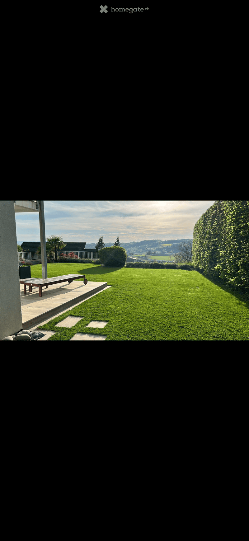 Well-maintained lawn with a wooden bench on a patio, surrounded by hedges and trees, with a scenic view of a valley and mountains in the background.
