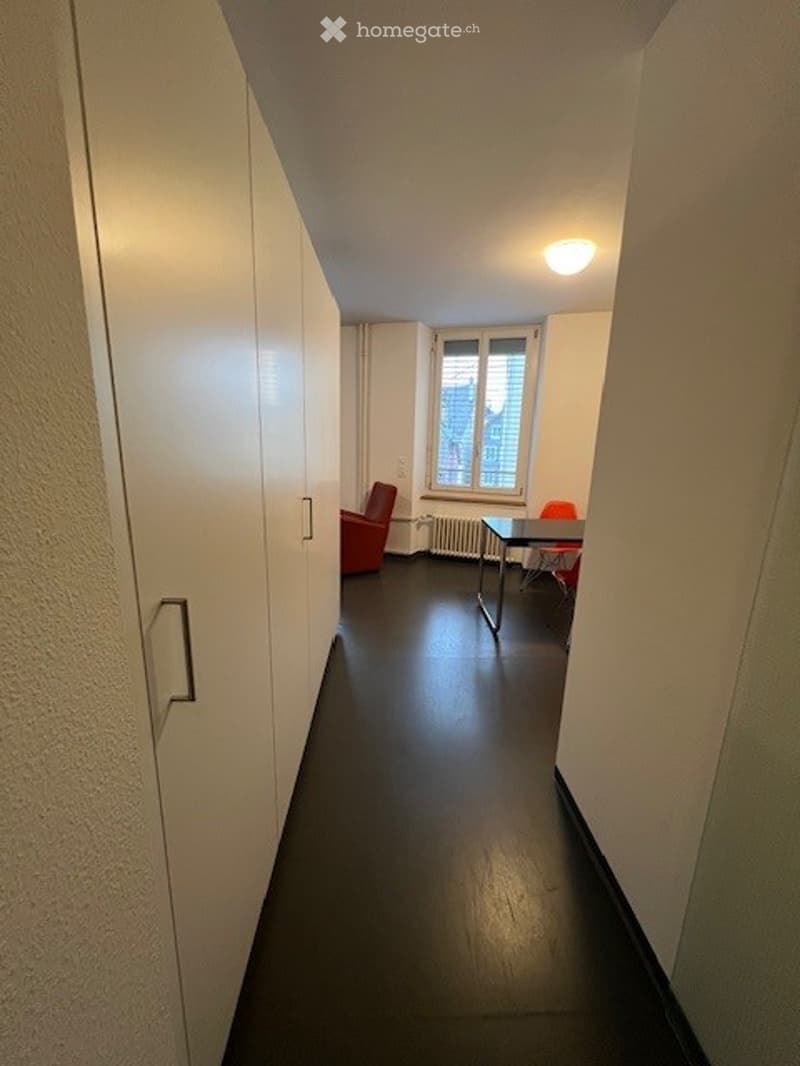 A narrow hallway with dark wooden flooring, white walls, and a small seating area featuring a red chair, a table, and an orange chair near a window with a radiator below.