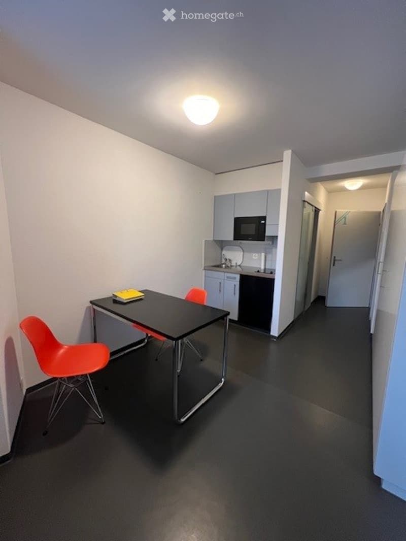 Modern, minimalist room with a black table, orange chairs, and a compact kitchen area featuring a microwave and cabinets.