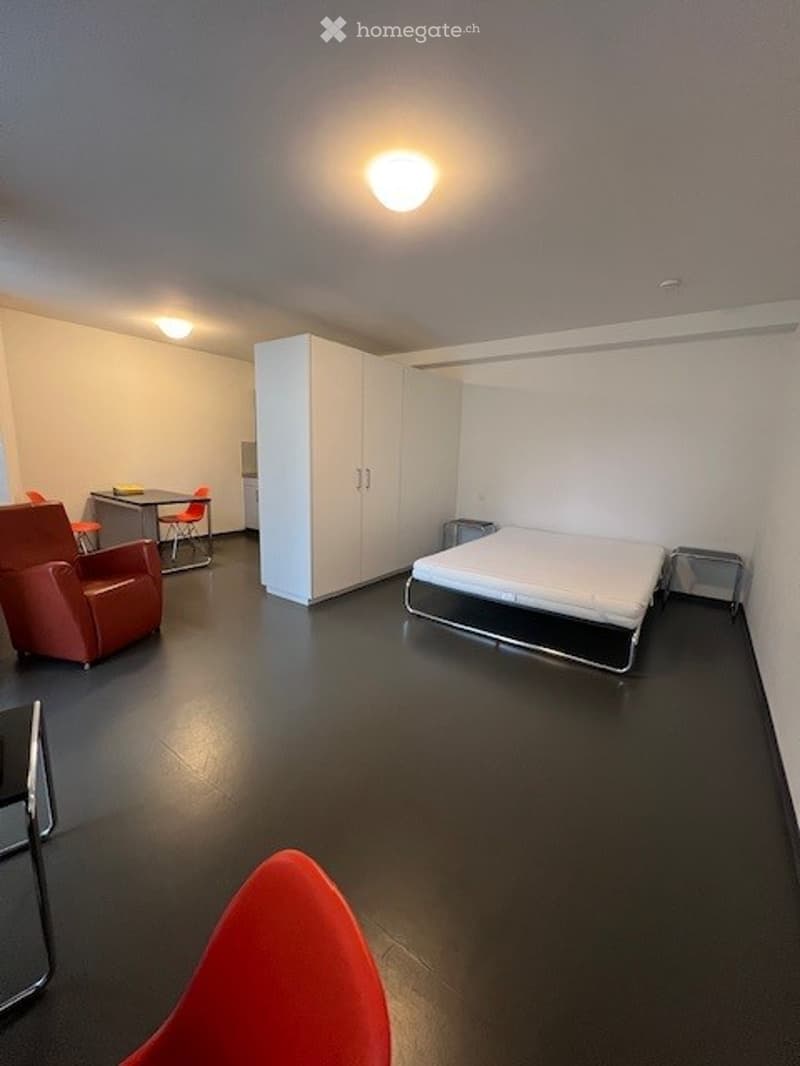 Modern, minimalist bedroom with a bed, red leather chair, small table, and built-in white wardrobe. The room features dark flooring and bright lighting.
