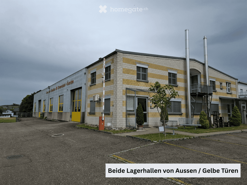 Two-story industrial building with yellow doors, tiled facade, and ventilation pipes. Small trees and benches are visible in front.