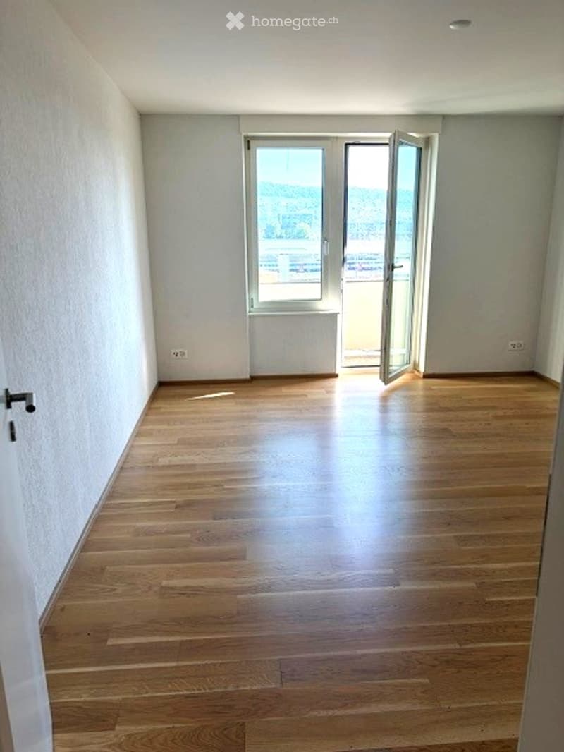 Empty room with wooden flooring, white walls, and a large window opening to a balcony.