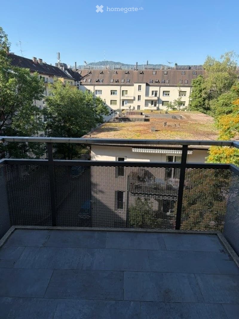 Balcony with metal railing, overlooking a cityscape with residential buildings and greenery.