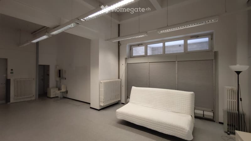 Minimalist room with a white sofa, a single chair, radiators, and a floor lamp. The room has large windows and a closed garage door.