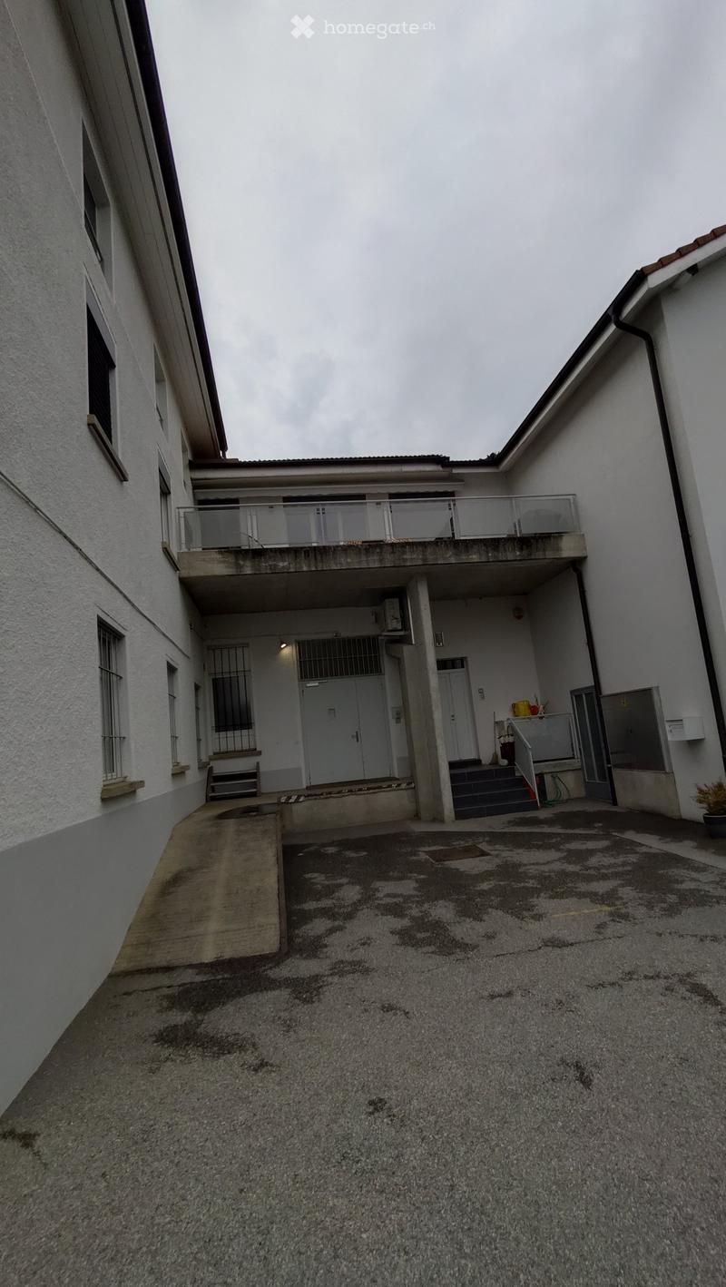 A multi-story building with a raised ground floor, featuring a balcony, stairs, and a concrete entrance area.