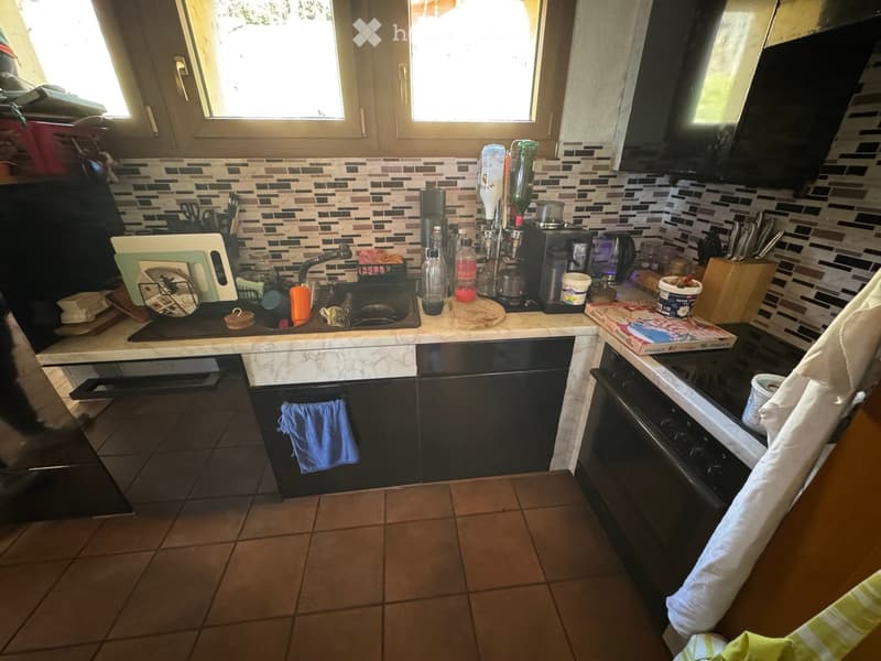 The kitchen features a marble countertop, a sink with a faucet, a coffee maker, a cutting board, a knife block, and various kitchen utensils. The cabinets are dark-colored, and the floor is tiled.