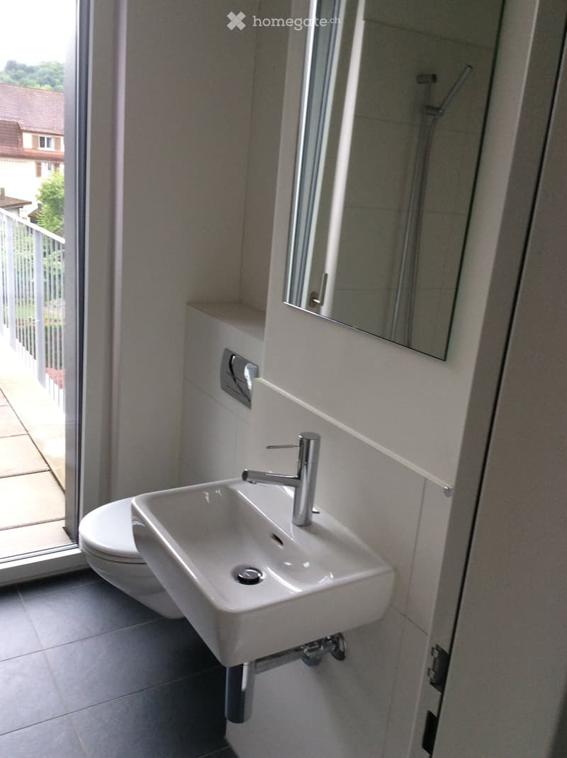 Modern bathroom with a white sink, chrome faucet, toilet, and mirror. The room has light-colored tiles and a large window offering a view of the outdoor area.