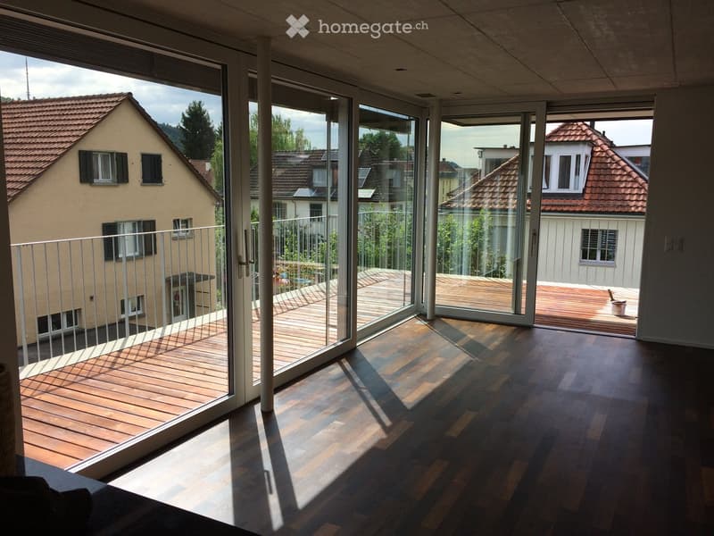 Spacious balcony with wooden decking, large glass sliding doors, and a metal railing, overlooking neighboring houses and greenery.