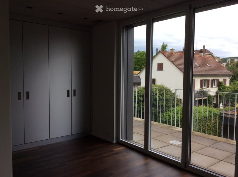 Modern interior with large sliding glass doors opening to a tiled balcony with metal railing, overlooking a residential neighborhood with houses and trees.