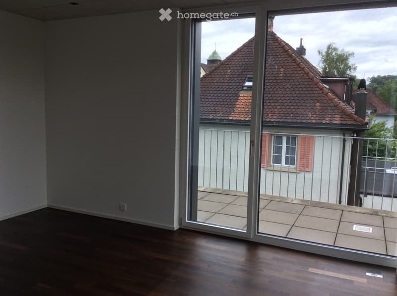 Empty room with dark wooden flooring and large sliding glass doors leading to a tiled balcony area with a view of neighboring rooftops.