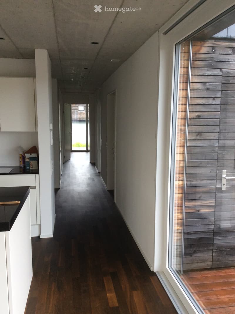A long, narrow hallway with wooden flooring, white walls, and a large window on the right side. The hallway leads to an open area with natural light and a view of the outdoors.