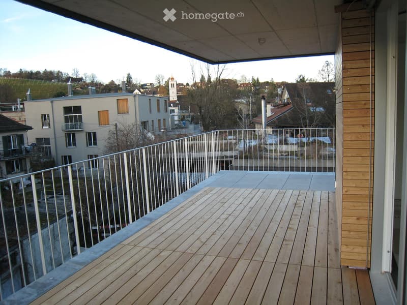 Wooden deck with metal railing, overlooking a town with houses, trees, and a church tower.