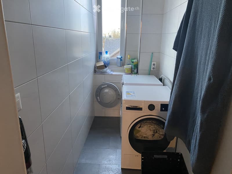 Bathroom with tiled walls, a washing machine, and a towel hanging on the wall. There is a window with various items on the sill.