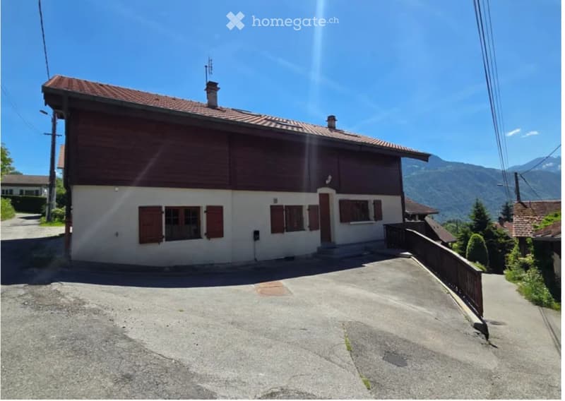 Two-story house with a red-tiled roof, white walls, and wooden shutters. The house features a chimney and is situated in a scenic mountainous area with a clear blue sky and surrounding greenery.