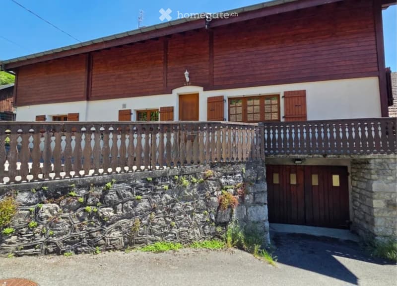 Two-story wooden house with white walls, multiple windows with shutters, and a stone retaining wall with decorative wooden railing.