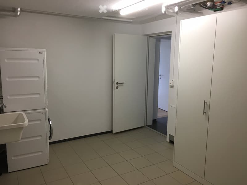 Empty room with tiled floor, white walls, a door leading to another space, and a washing machine with a sink.