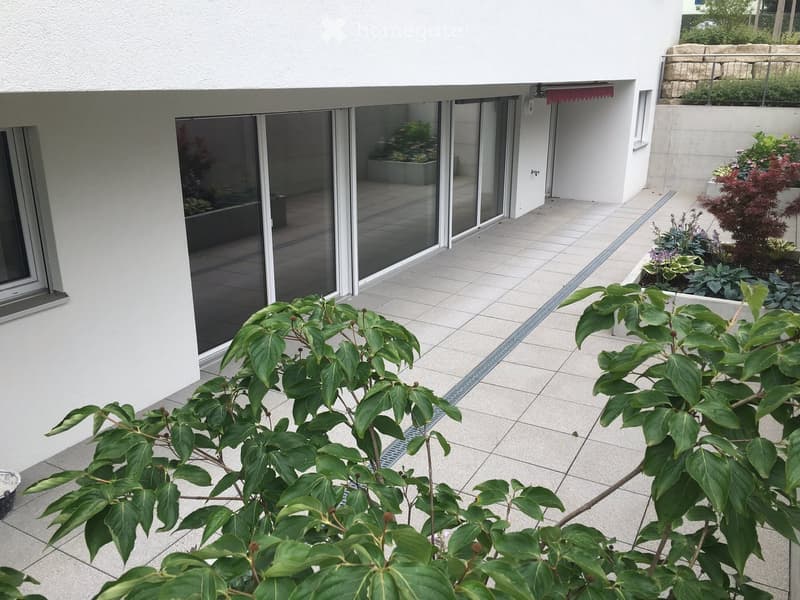 A modern patio area with tiled flooring, large sliding glass doors leading to an indoor space, and various potted plants and shrubs.