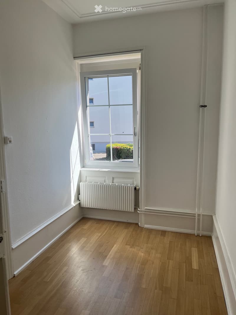 Empty room with wooden flooring, white walls, a radiator, and a large window with a view of the outside.