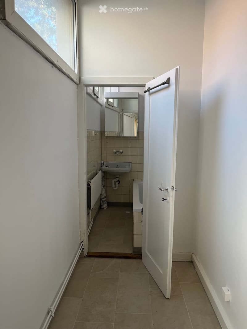 A narrow hallway with tiled flooring and white walls, leading to an open bathroom door. The bathroom contains a sink, mirror, and radiator.