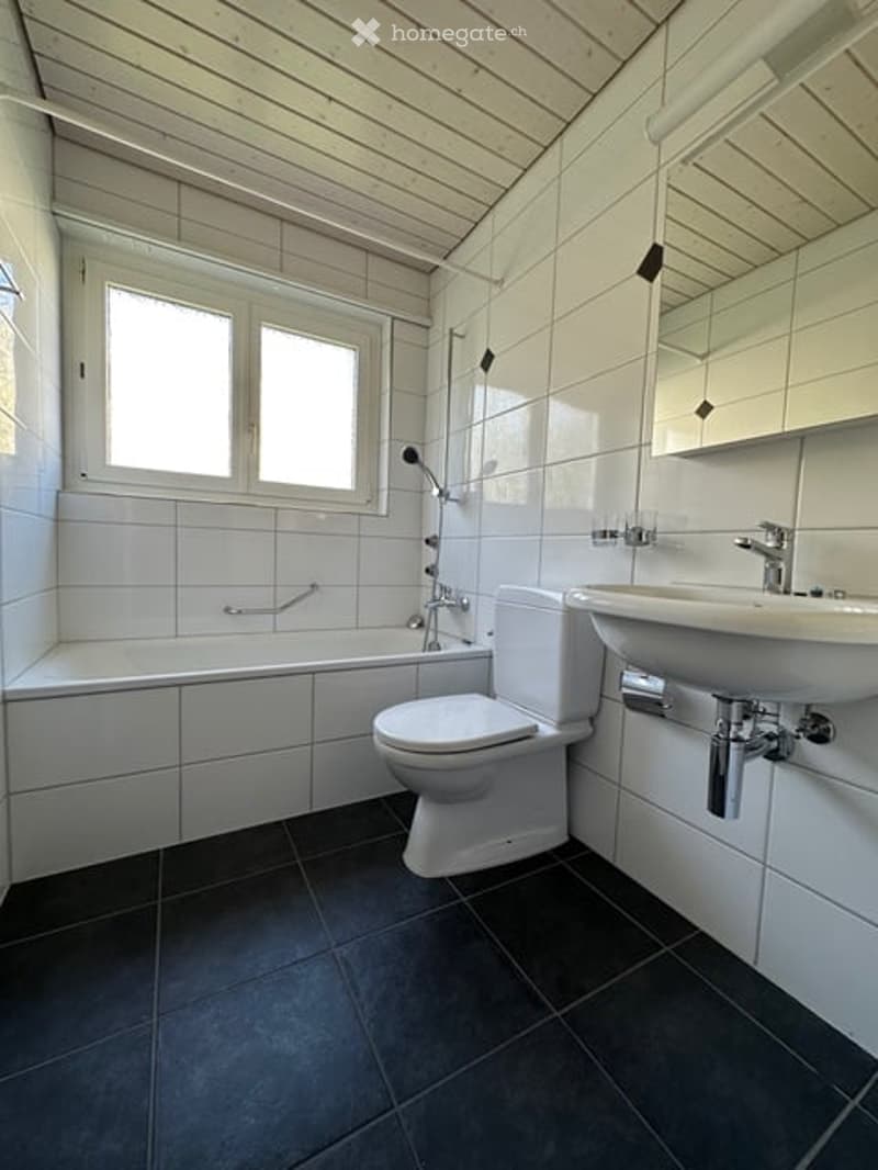Modern bathroom with white tiles, dark grey floor, bathtub, toilet, sink, and mirror.