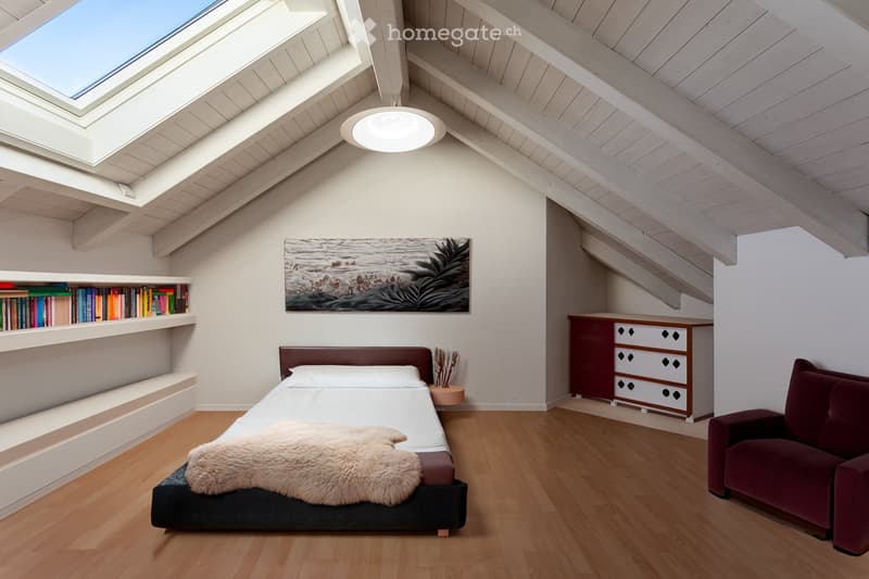 Modern attic bedroom with wooden floors, slanted ceilings, skylight windows, a bed with a fur throw, a red sofa, built-in bookshelf, and a picture frame on the wall.