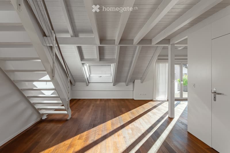 Bright, airy hallway with wooden flooring, white walls, and exposed wooden beams on the ceiling. Features a staircase leading upwards and a large window allowing natural light to flood the space.