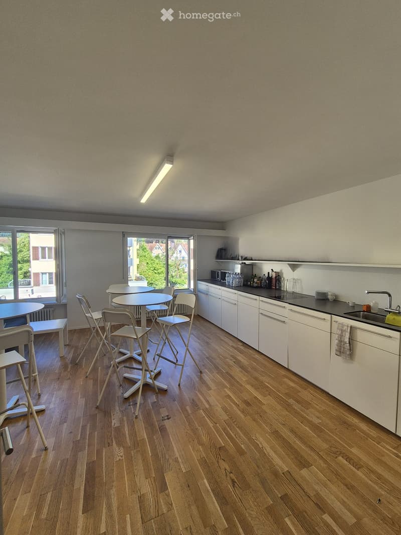 Modern kitchen with white cabinets, wooden flooring, and a dining area with a round table and chairs. The kitchen features a sink, microwave, and various small appliances on the counter.