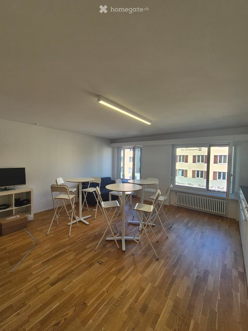 A furnished room with wooden flooring, several tables and chairs, a sofa, and a TV on a shelf.