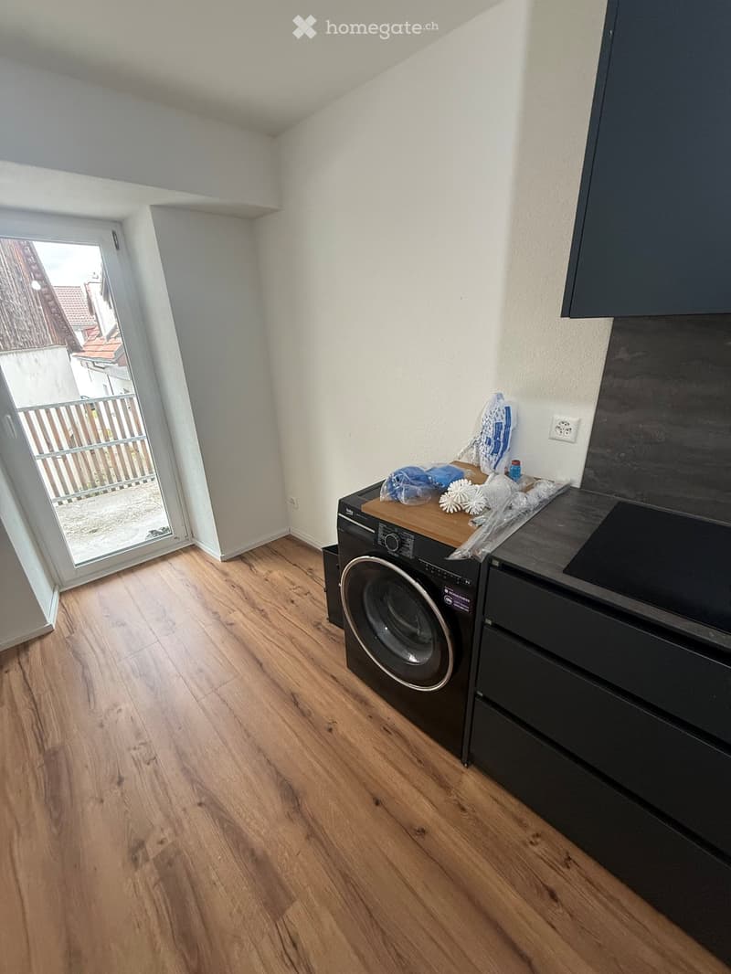 A small room with wooden flooring, a washing machine, a dishwasher, and a small table with some items on it. There is a sliding glass door leading to a balcony.