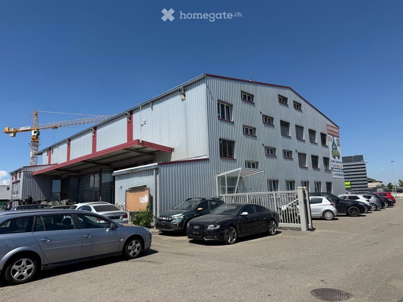 A large industrial-style building with a metal facade and red trim, surrounded by a parking lot with several cars.