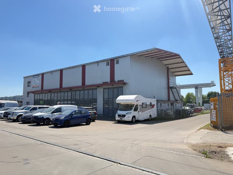 A modern commercial building with a large parking lot, featuring several parked vehicles including cars and a camper van.