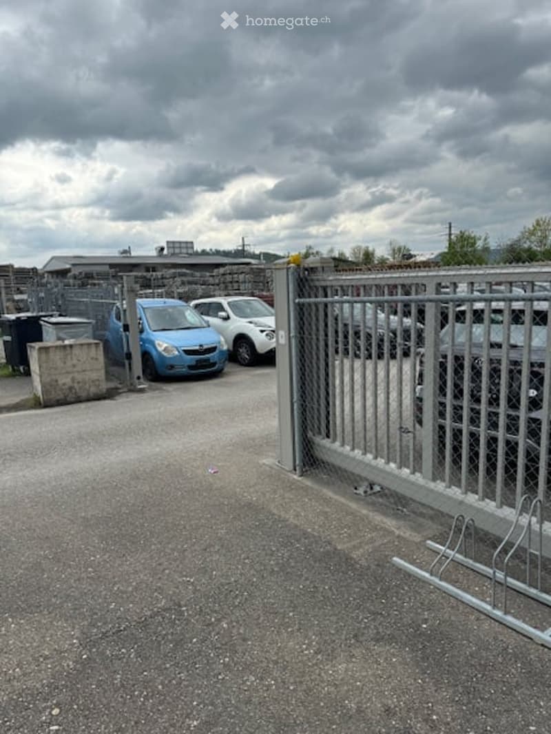 Open parking area with metal gate, paved ground, multiple parked cars, trash bins