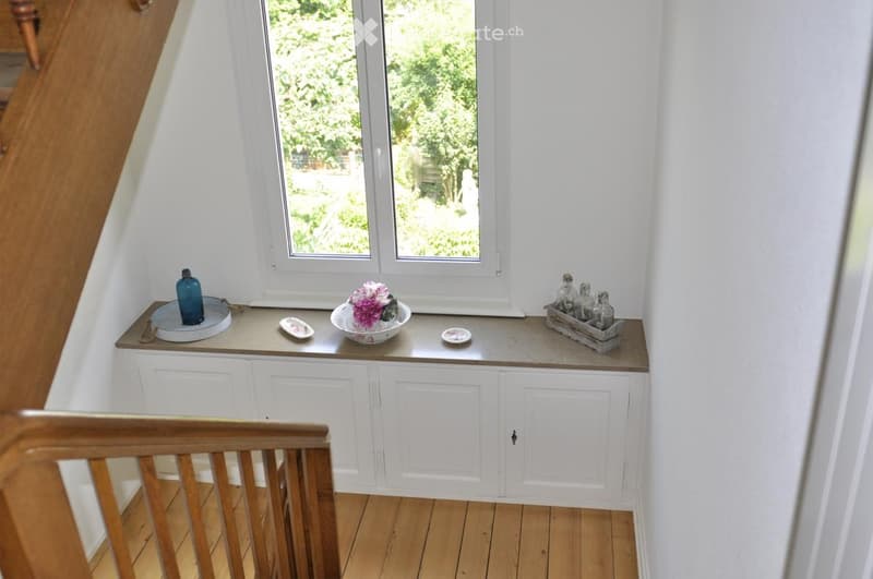 A narrow hallway with white walls, wooden flooring, and built-in white cabinets beneath a large window. Decorative items, including a vase with flowers and small plates, are placed on the windowsill.