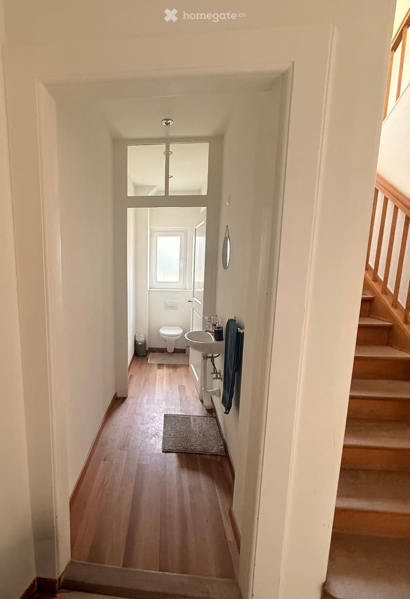 A narrow hallway with wooden flooring, leading to a small bathroom and a staircase. The bathroom contains a toilet, sink, and a small window. The hallway has white walls and a small rug placed near the bathroom entrance.