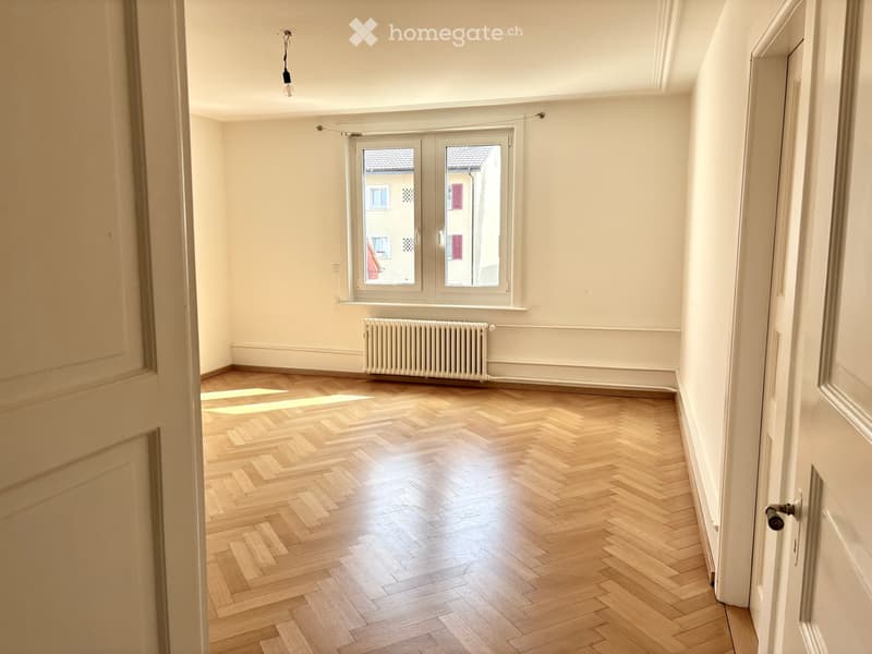 Empty room with wooden herringbone flooring, white walls, and a radiator under a large window. Natural light streams in, illuminating the space.