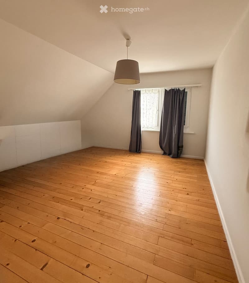 Empty room with wooden flooring, white walls, and a single window with dark curtains. A simple pendant light hangs from the ceiling.