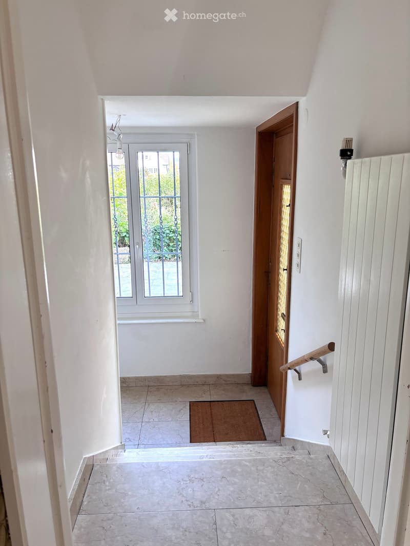 A clean, well-lit hallway with white walls, tiled floors, and a wooden door leading to another room. A small doormat is placed near the door, and a railing is visible on the right side.