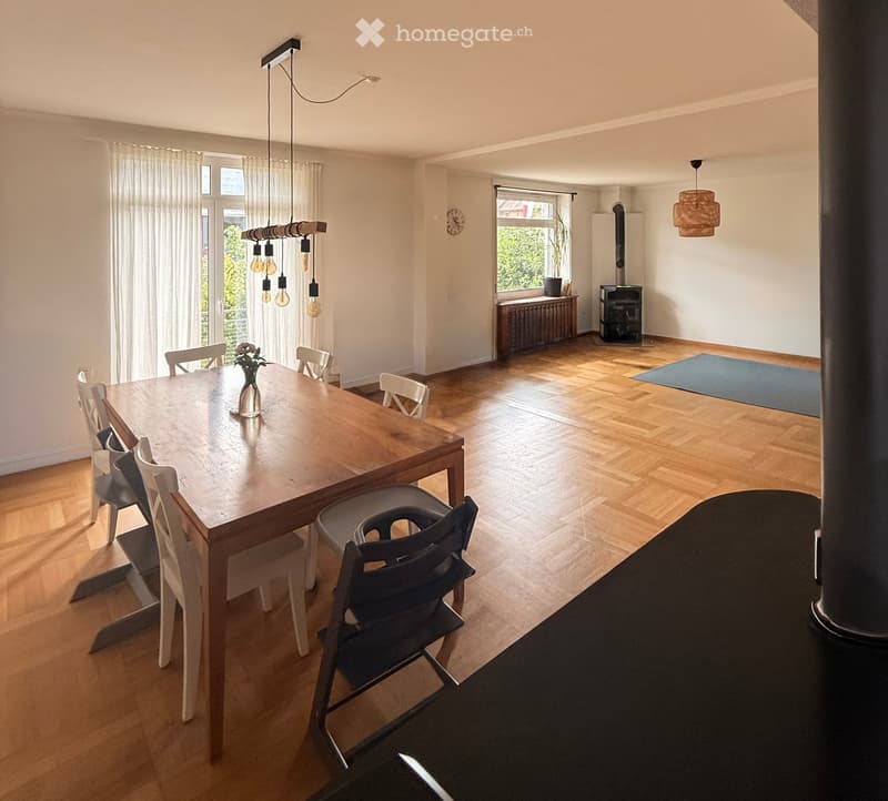 Bright dining area with wooden dining table, chairs, vase, pendant lights, fireplace, radiator, and large windows with curtains.