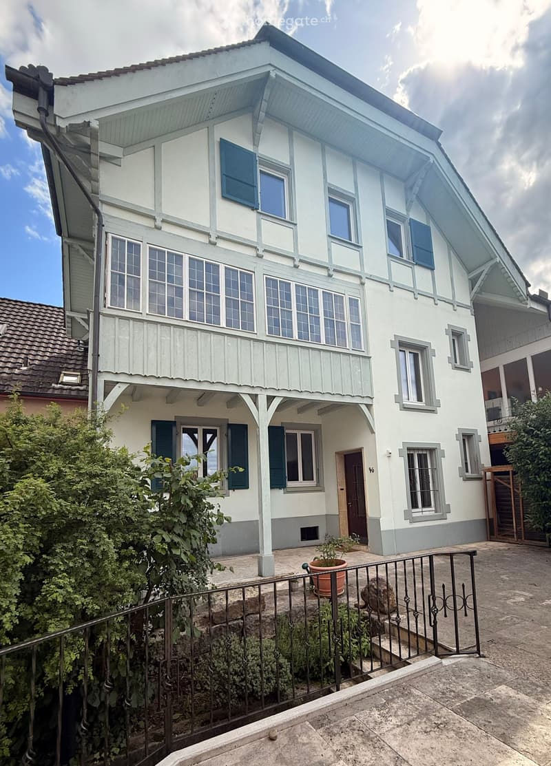 Two-story house with white and light blue exterior, multiple windows with shutters, and a small garden area with a potted plant and a metal fence.