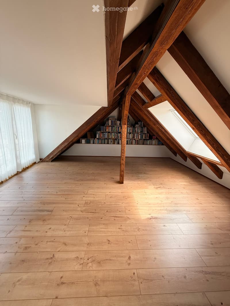 An attic space with wooden beams and a skylight, featuring a wooden floor and built-in bookshelves along one wall.