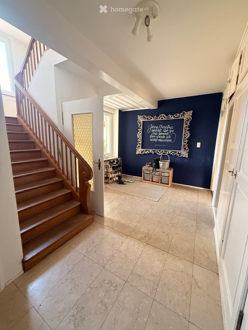 Spacious hallway with wooden staircase, tiled floor, and a blue accent wall featuring a framed quote. Storage unit and shoe rack visible in the background.