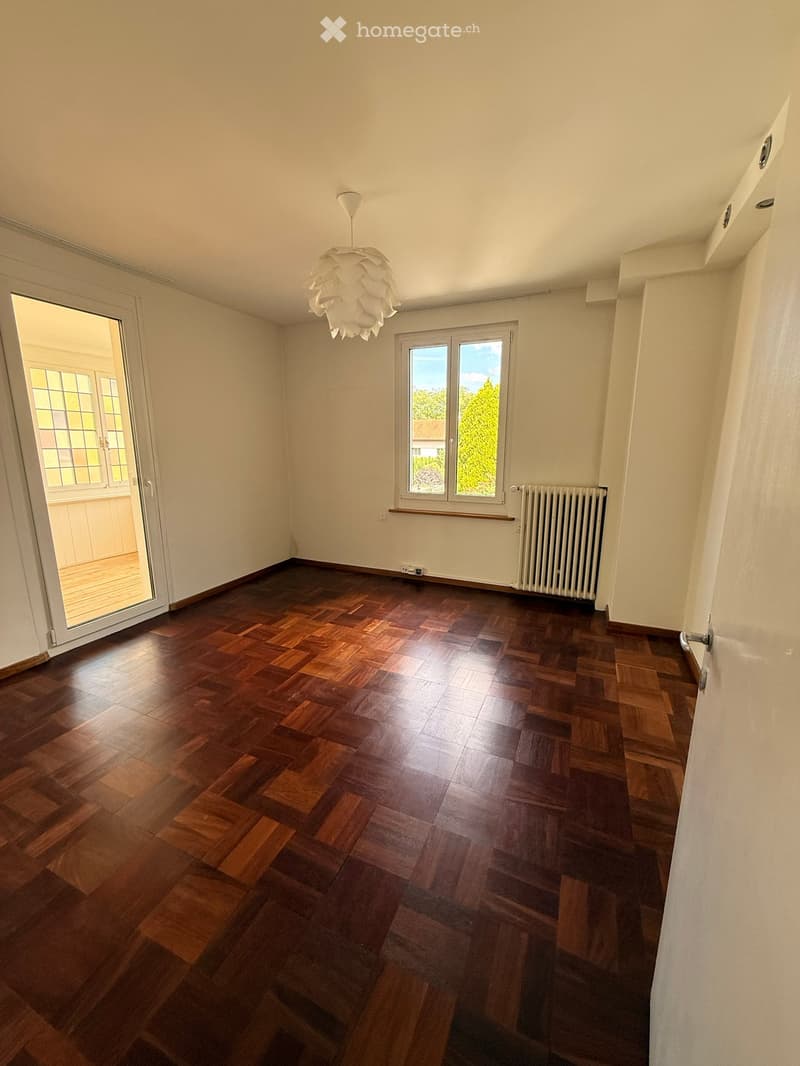 Empty room with wooden flooring, white walls, and large windows. Features a radiator and a modern hanging light fixture.