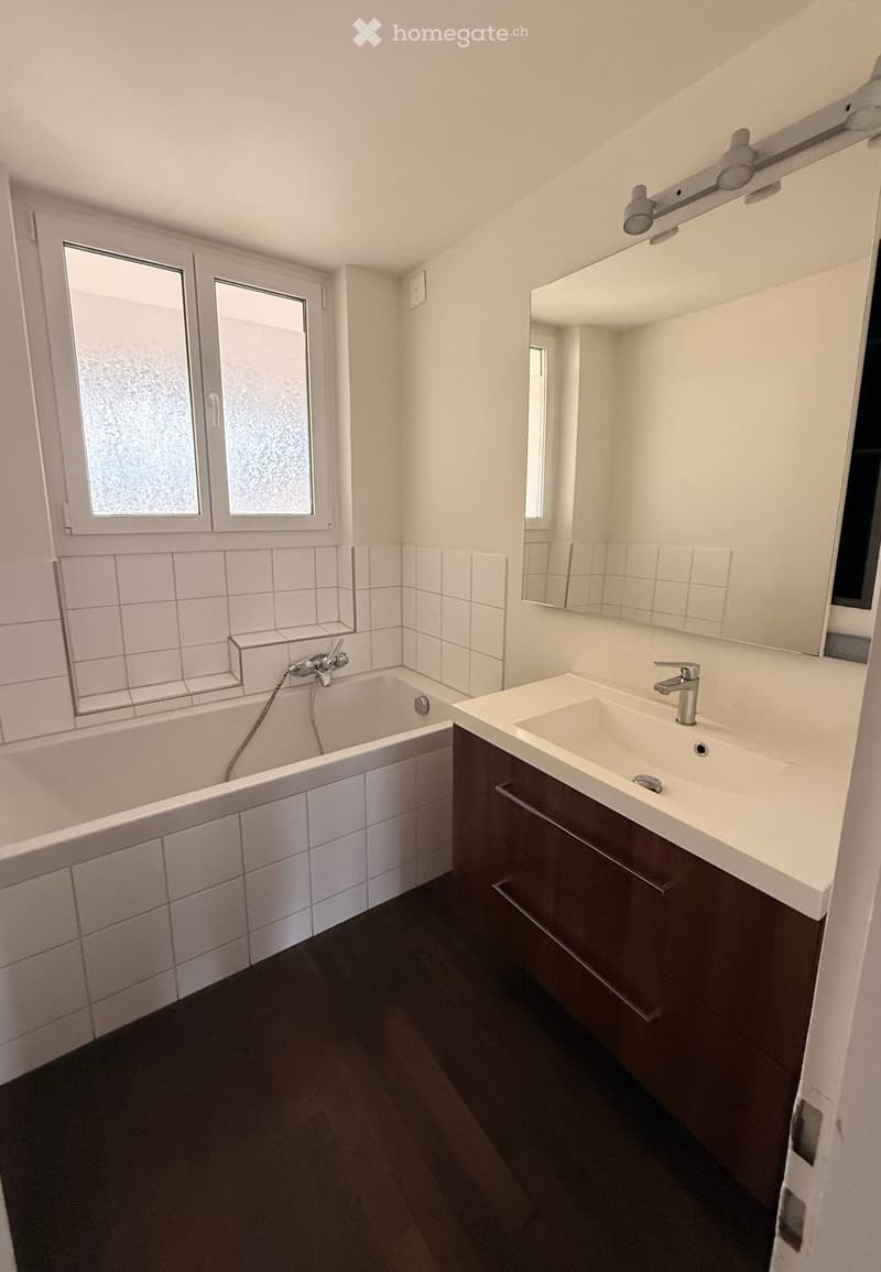 Modern bathroom with a white tiled bathtub, a white sink with a chrome faucet, a large mirror, and a dark wooden cabinet. The walls are tiled in white, and the floor is dark.