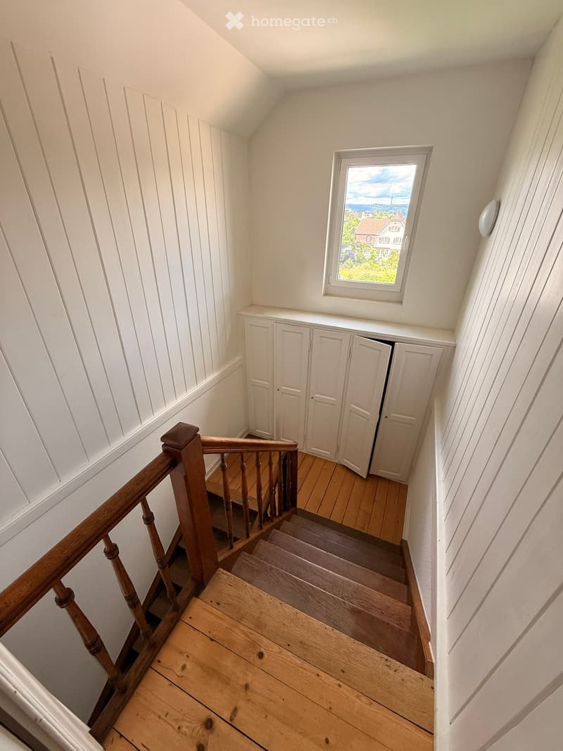 Wooden staircase with a wooden handrail and balusters, white paneled walls, a small window at the top showing a view of the outside, and built-in white cabinets on one side.