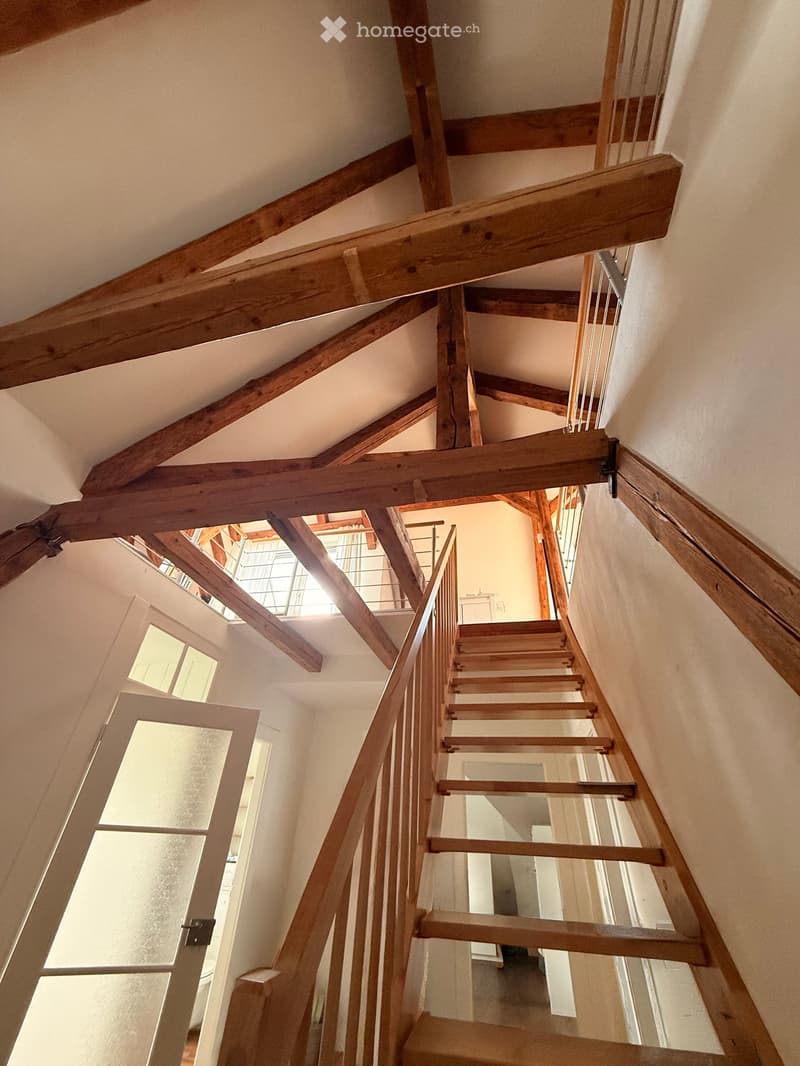 Wooden staircase with exposed wooden beams and a modern glass railing, leading to an upper level with visible windows and doors.
