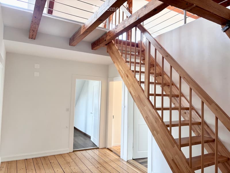 Bright hallway with wooden flooring, white walls, and a wooden staircase leading upwards. Features exposed wooden beams and modern lighting fixtures.